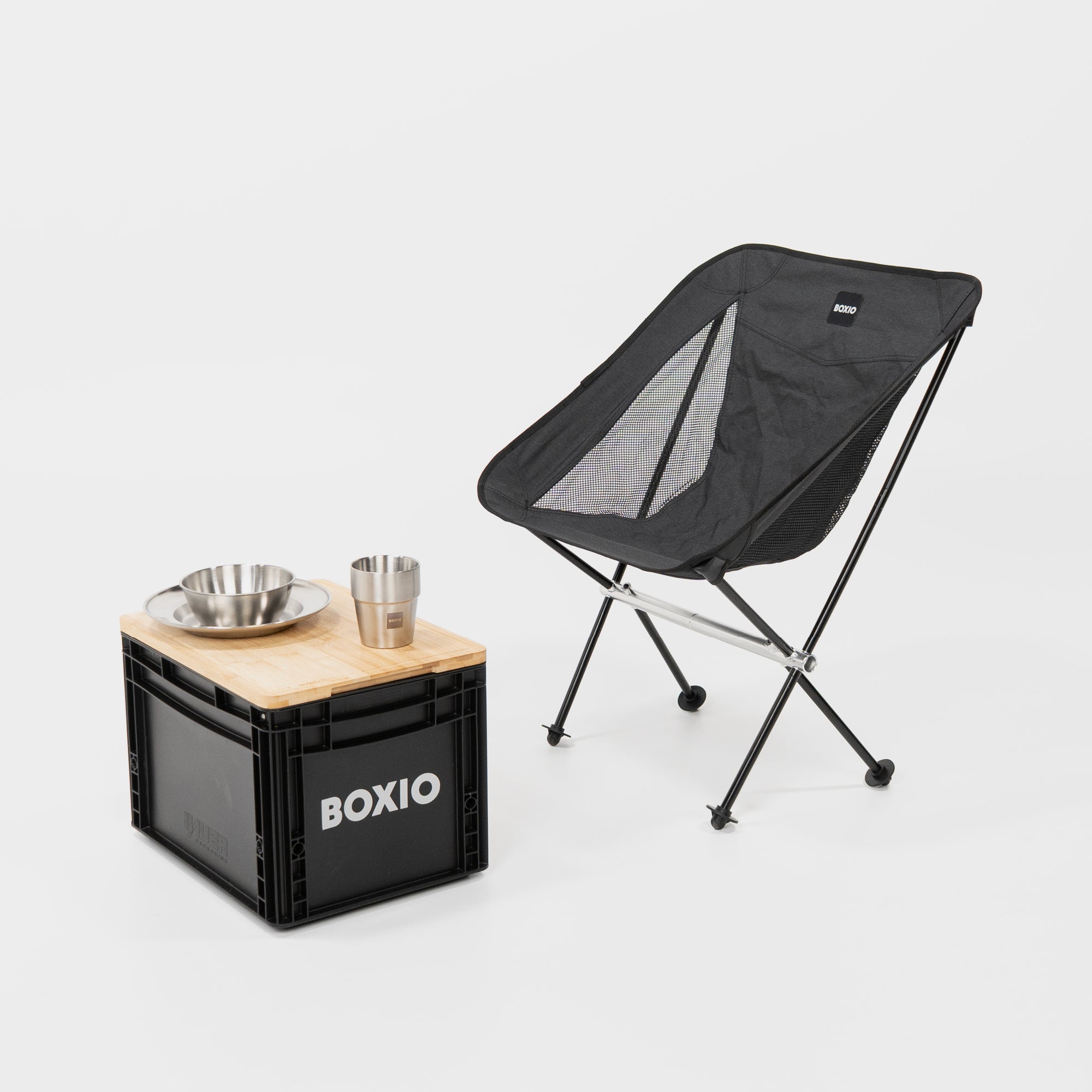 Campingstuhl mit seitlichen Lüftungsschlitzen im Bezug. Davor eine BOXIO Eurobox mit Schneidebrett auf dem Deckel. Darauf das BOXIO Campinggeschirr vor weißem Hintergrund