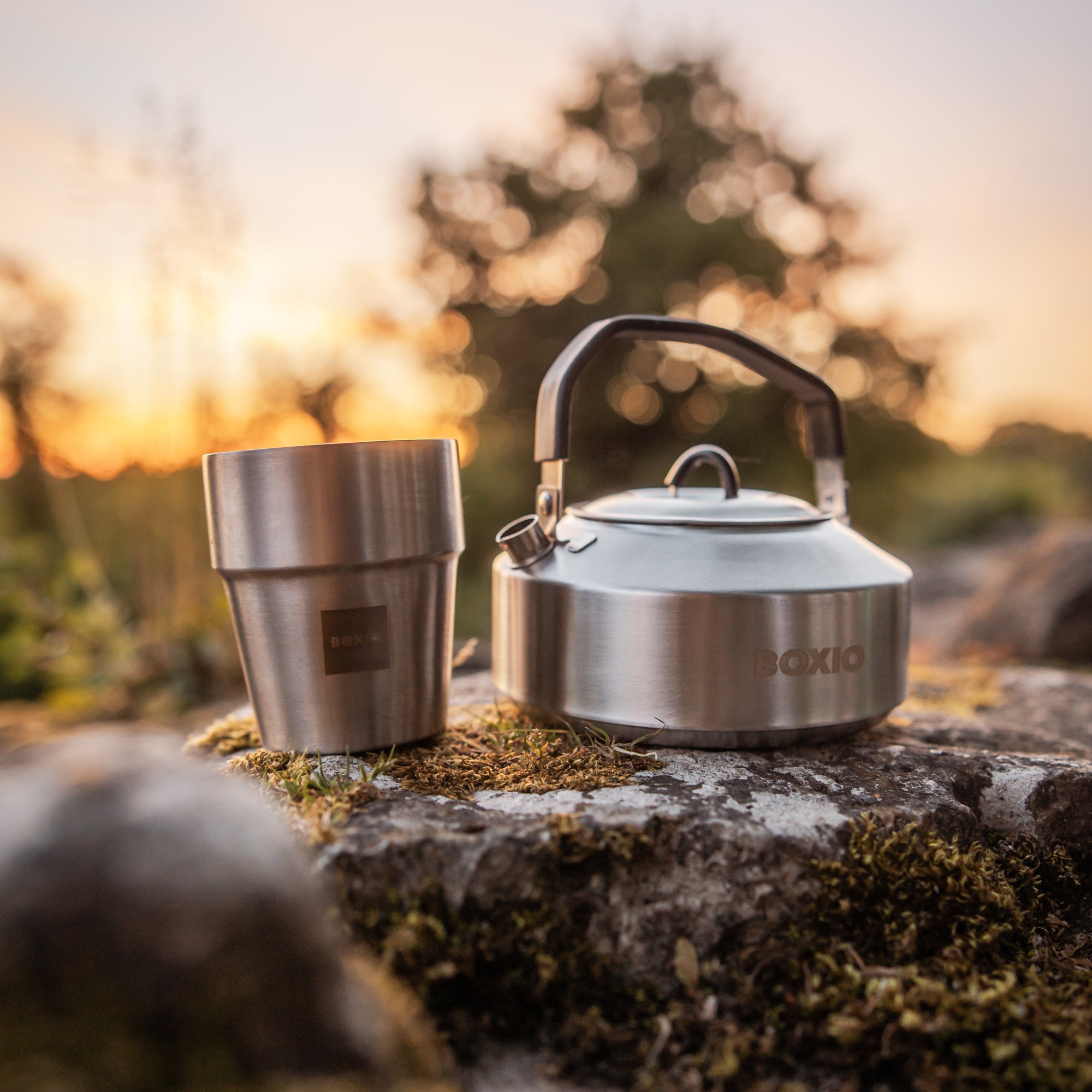 Waldhintergrund auf einem Stein steht ein Wasserkessel aus Edelstahl und ein Trinkbecher aus Edelstahl