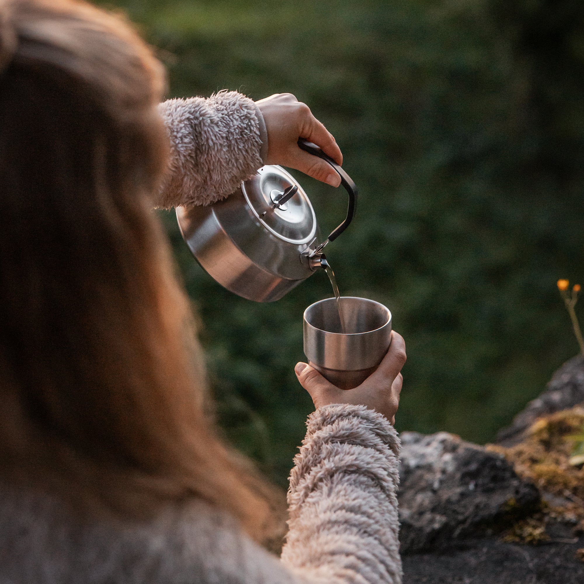 Waldszenerie, Frau gießt aus Wasserkessel Flüssigkeit in einen Edelstahlbecher