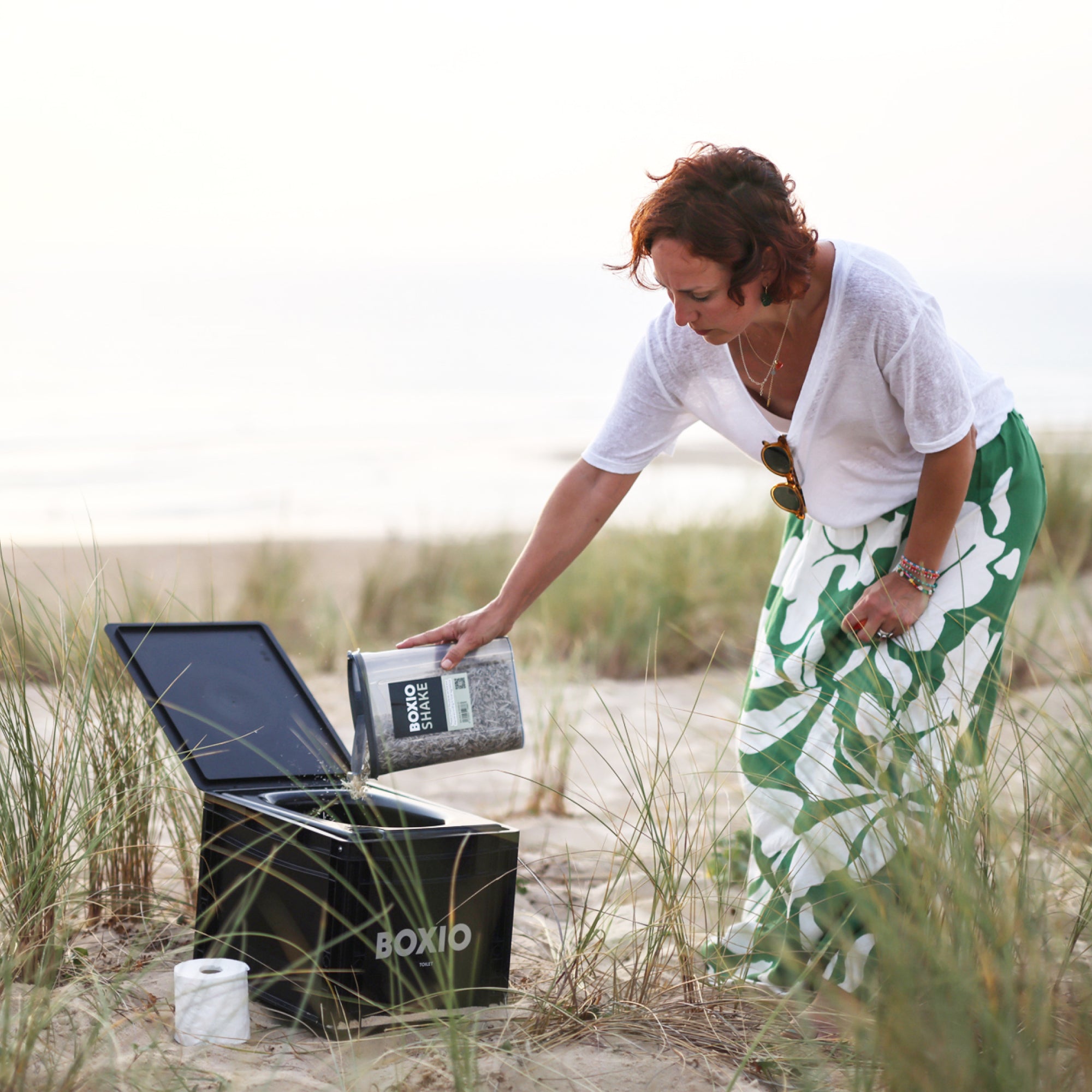 Frau in Strandkleidung gießt eine Mischung in eine schwarze Box auf einem Sandstrand mit Gras im Hintergrund.