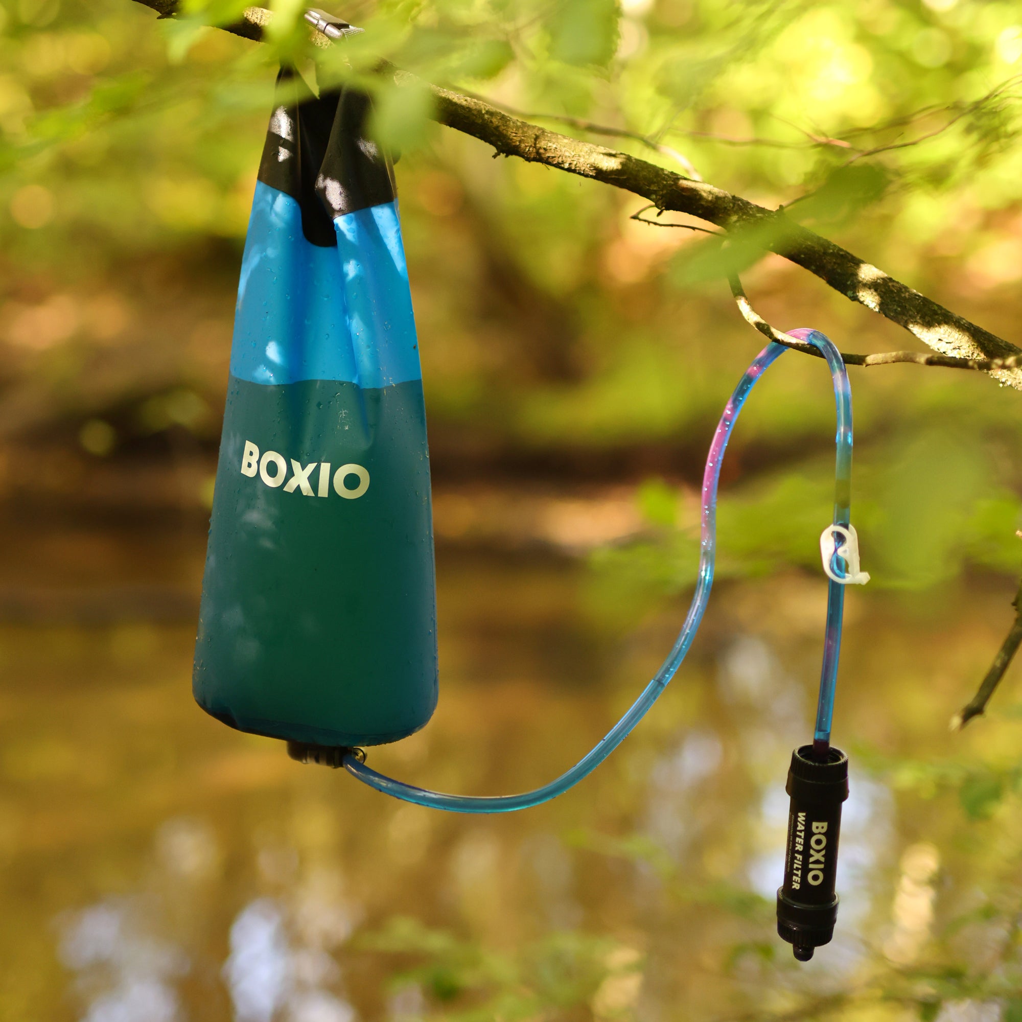 Hängende Wasserfilter-Flasche von BOXIO mit Schlauch, im Hintergrund ein fließendes Gewässer und grünes Laub.