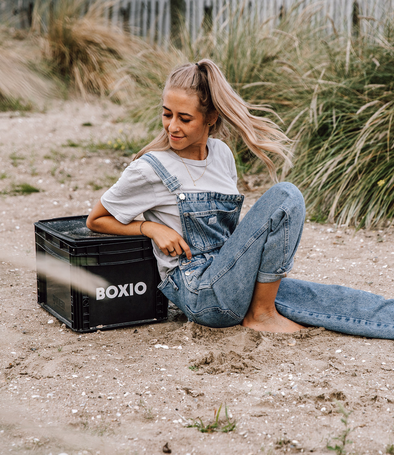 Junge Frau sitzt im Sand, trägt eine blaue Latzhose und ein T-Shirt, neben ihr steht eine schwarze Box mit dem Schriftzug "BOXIO".