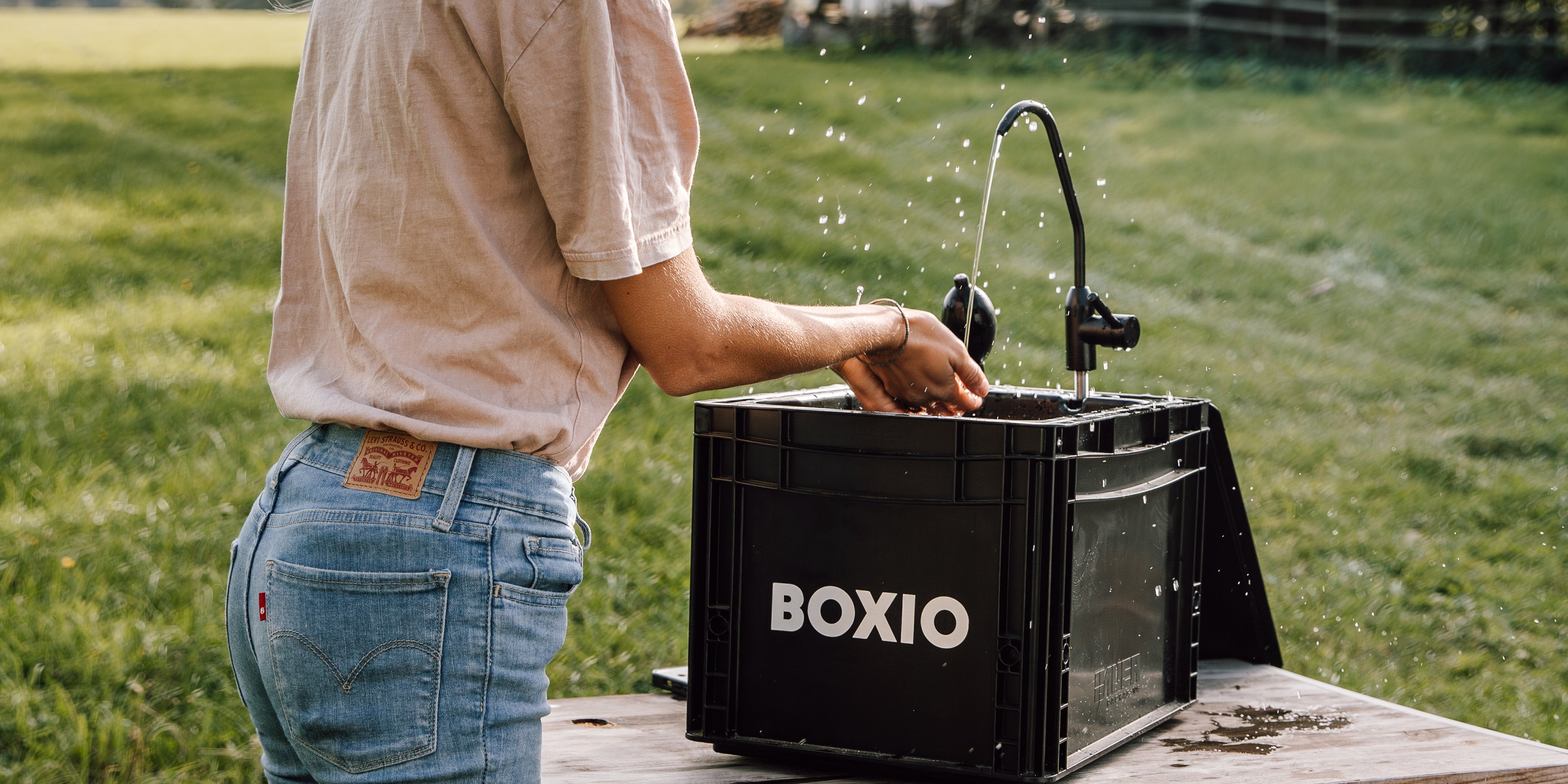Person mit blonden Haaren, die Hände an einem mobilen Wasserhahn in einem Garten wäscht.
