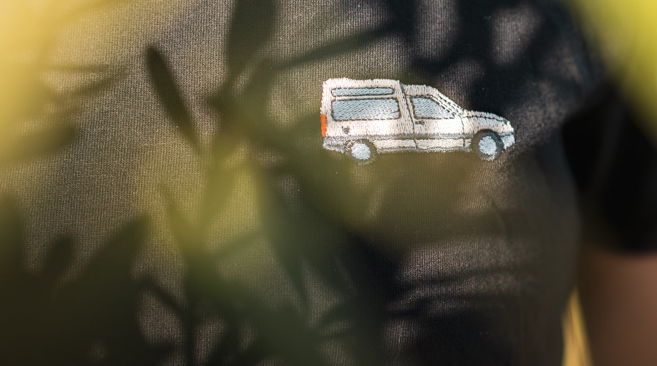 Schwarzes T-Shirt mit aufgesticktem, grafischen Van-Motiv, umgeben von sanftem Pflanzenlicht.