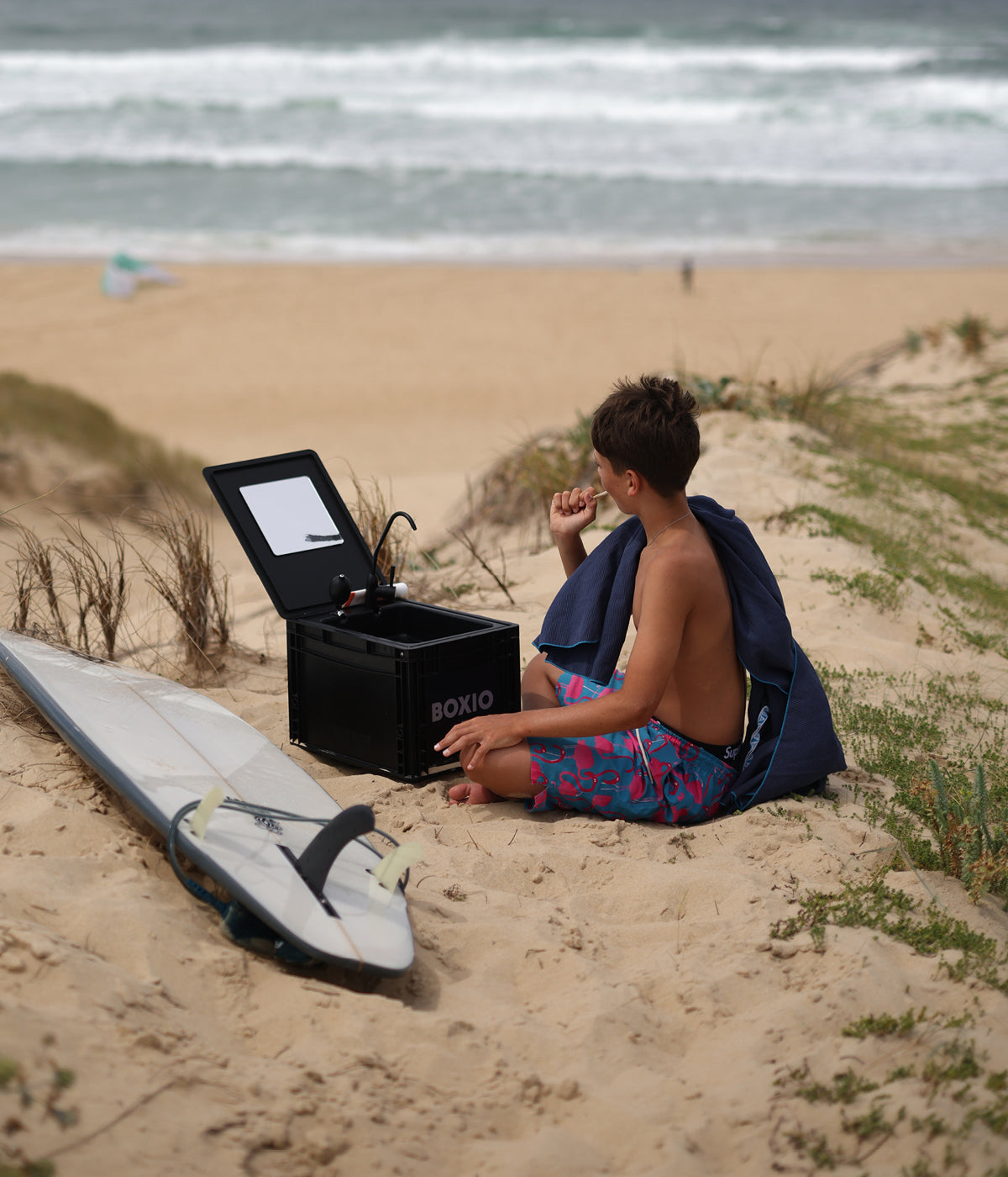 Junge sitzt im Sand putzt sich die Zähne mit der Boxio Wash und eiN Surfboard liegt neben ihm 